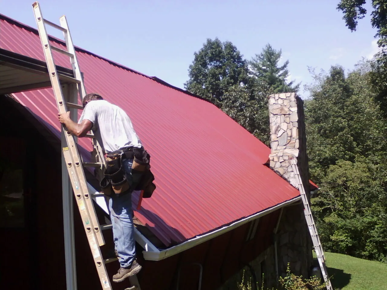 Roofer on a ladder beside a red metal roof and stone chimney — Soffit & Fascia Repair by Roofing Brothers in Eglin Afb